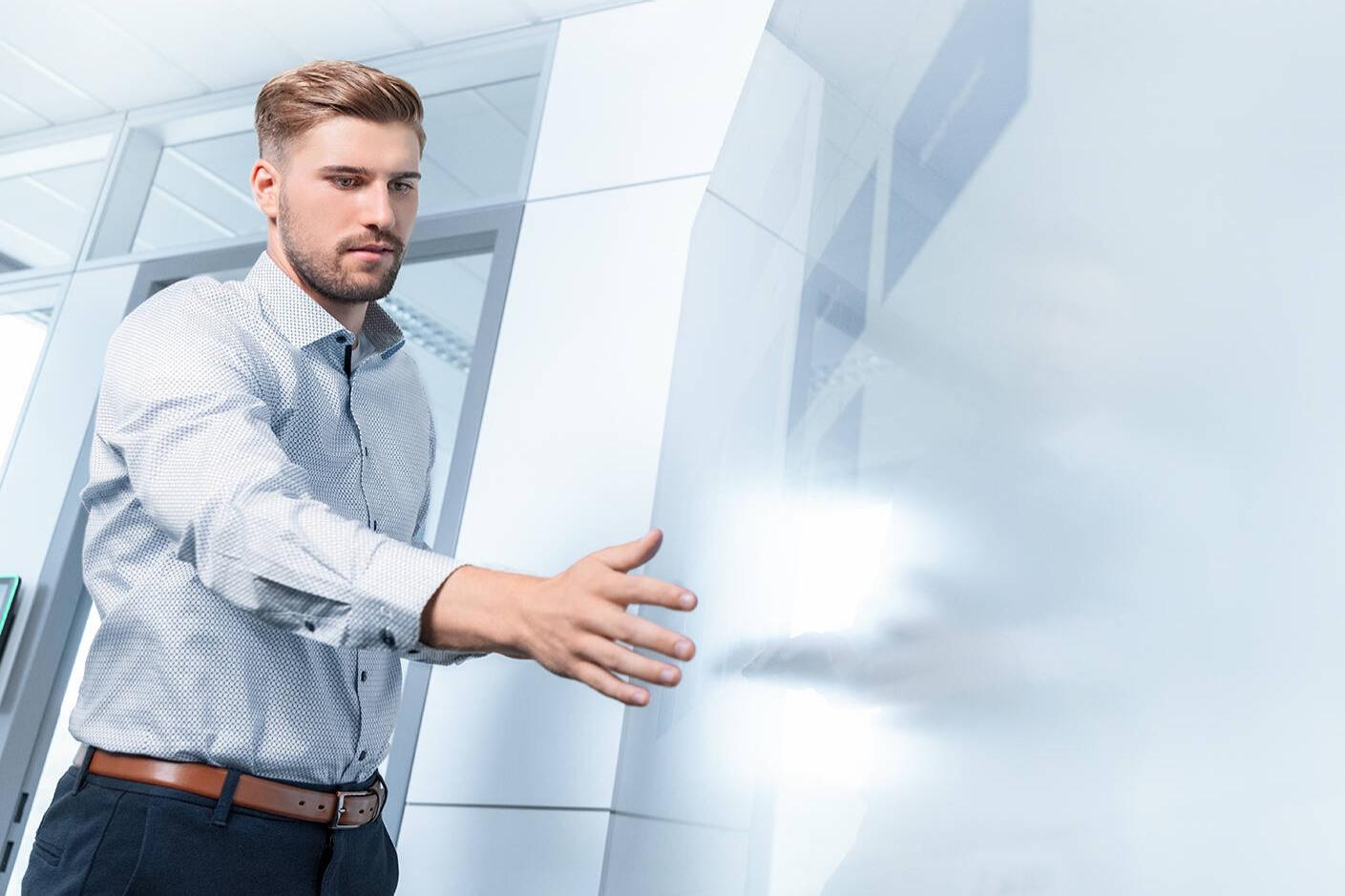 Fabian blickt auf einen Touchmonitor und berührt diesen mit seiner rechten Hand. Er trägt ein helles gemustertes Hemd und befindet sich in einer modernen Büroumgebung.
