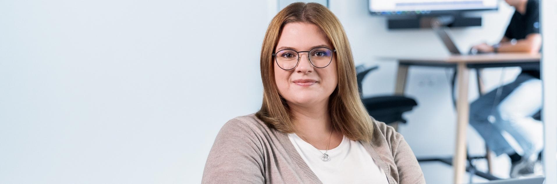 Eine weibliche Person sitzt schreibend an einem Laptop und blickt freundlich in die Kamera
