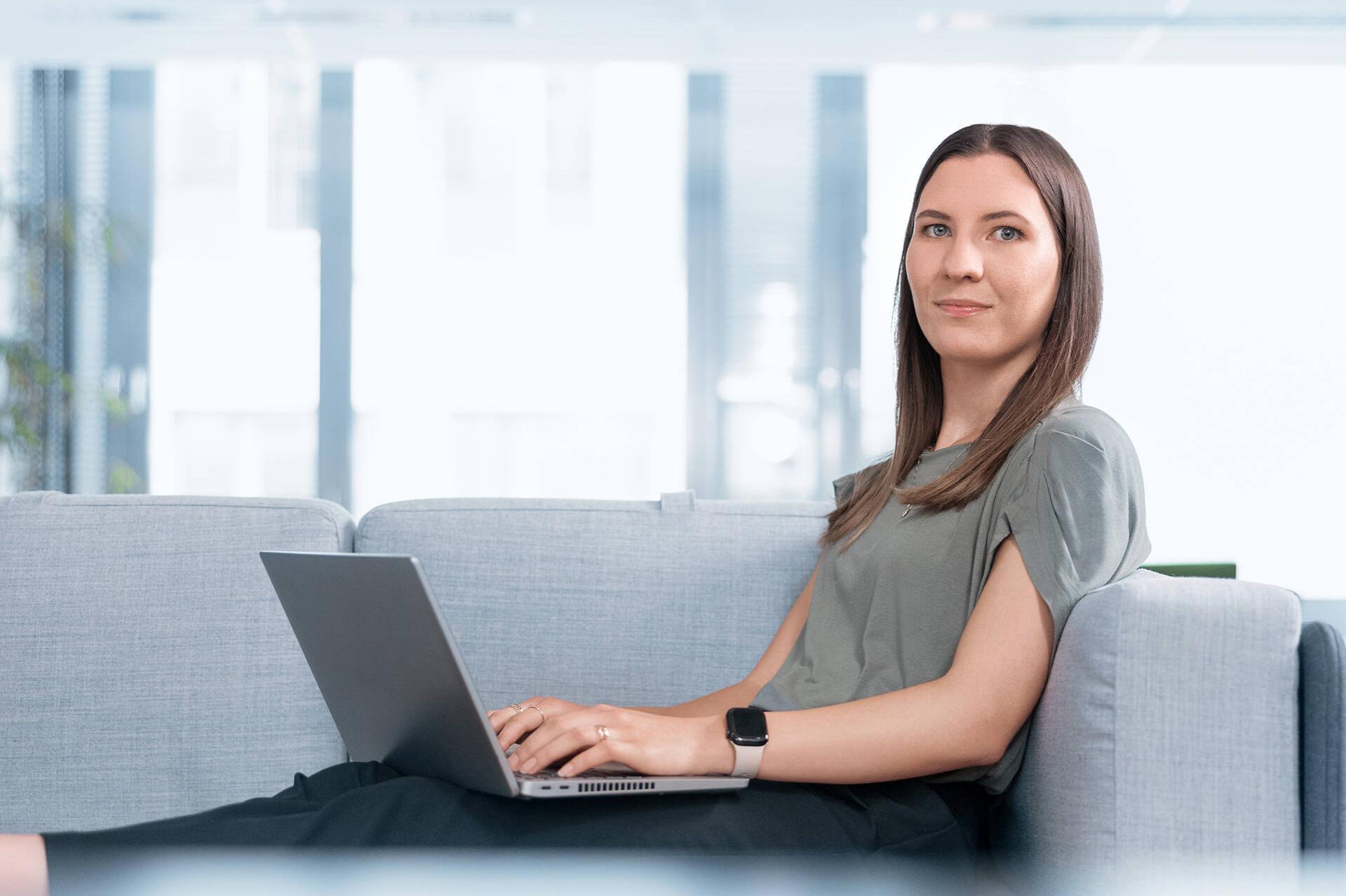 Eine weibliche Person sitzt schreibend an einem Laptop und blickt freundlich in die Kamera