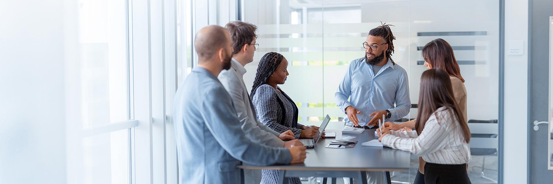 Six people stand around a desk in a bright, modern office and look at a person in the center. This man has the floor and is talking to his female colleague on his right, while the others make notes or just listen.