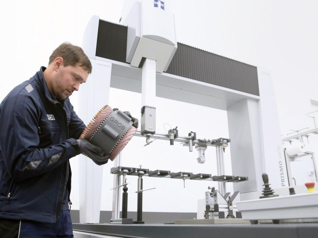 A technician in a ZEISS facility inspects a mechanical component in front of a ZEISS coordinate measuring machine.