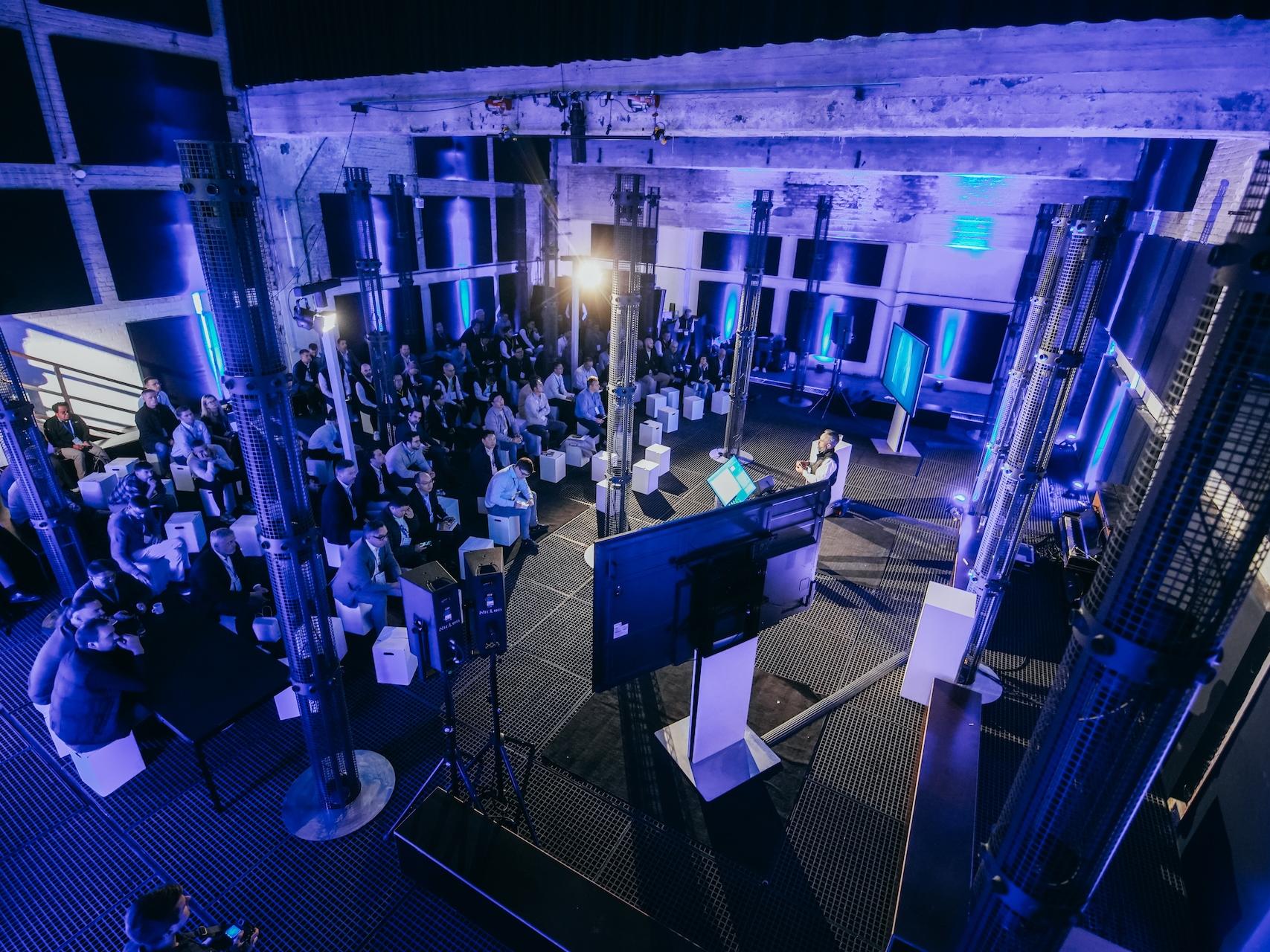 Audience attending a ZEISS presentation or event in a modern, industrial-style venue with stage lighting and large display screens.