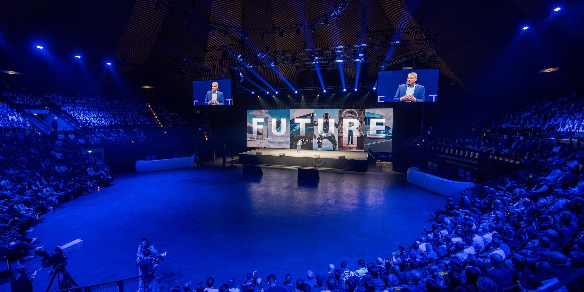 A large audience watches a ZEISS event in a circular auditorium with a prominent stage displaying the word "FUTURE" and speakers under dramatic blue lighting.