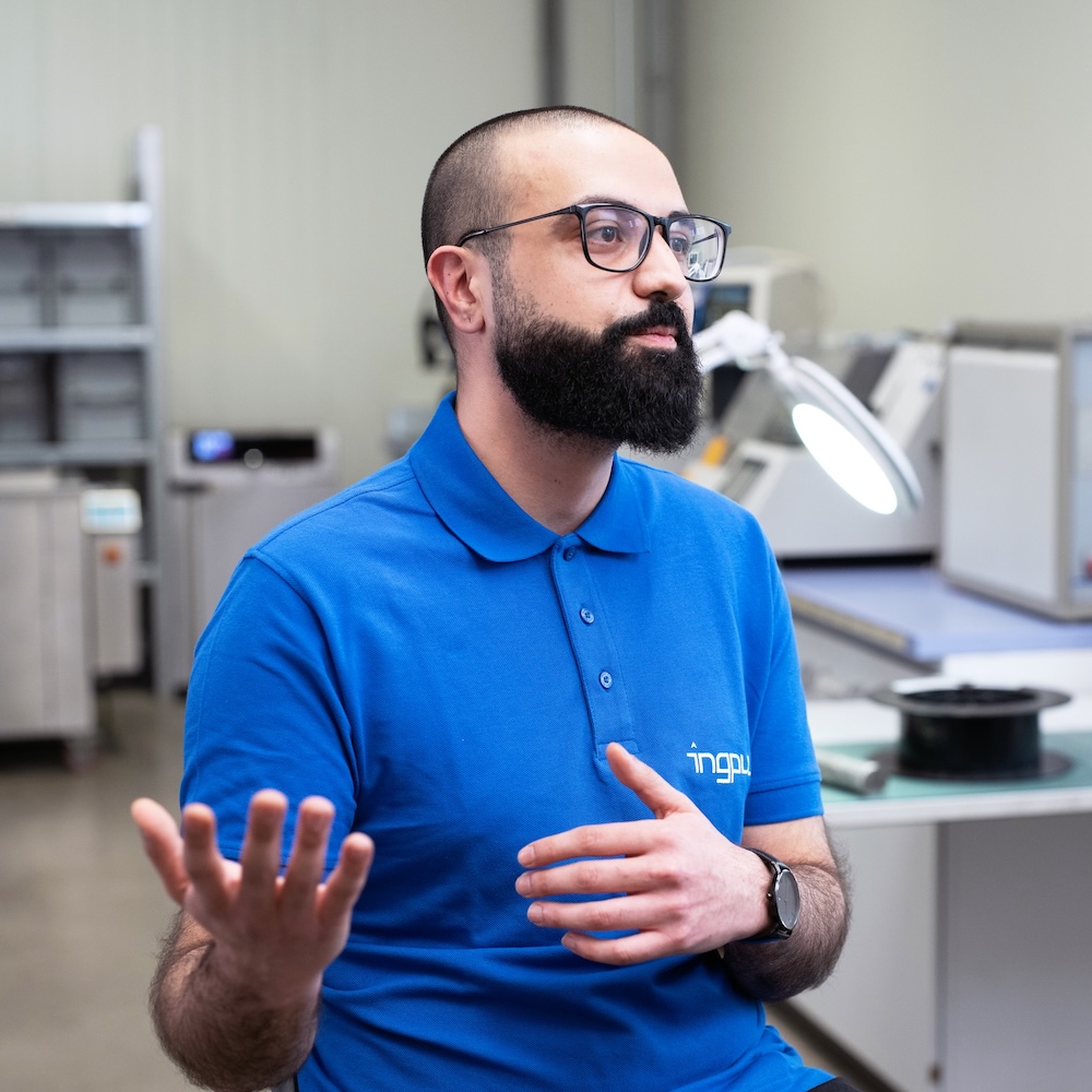 Ali El-Toufaili wearing glasses and a blue polo shirt is speaking in a modern lab equipped with ZEISS inspection and measurement instruments.