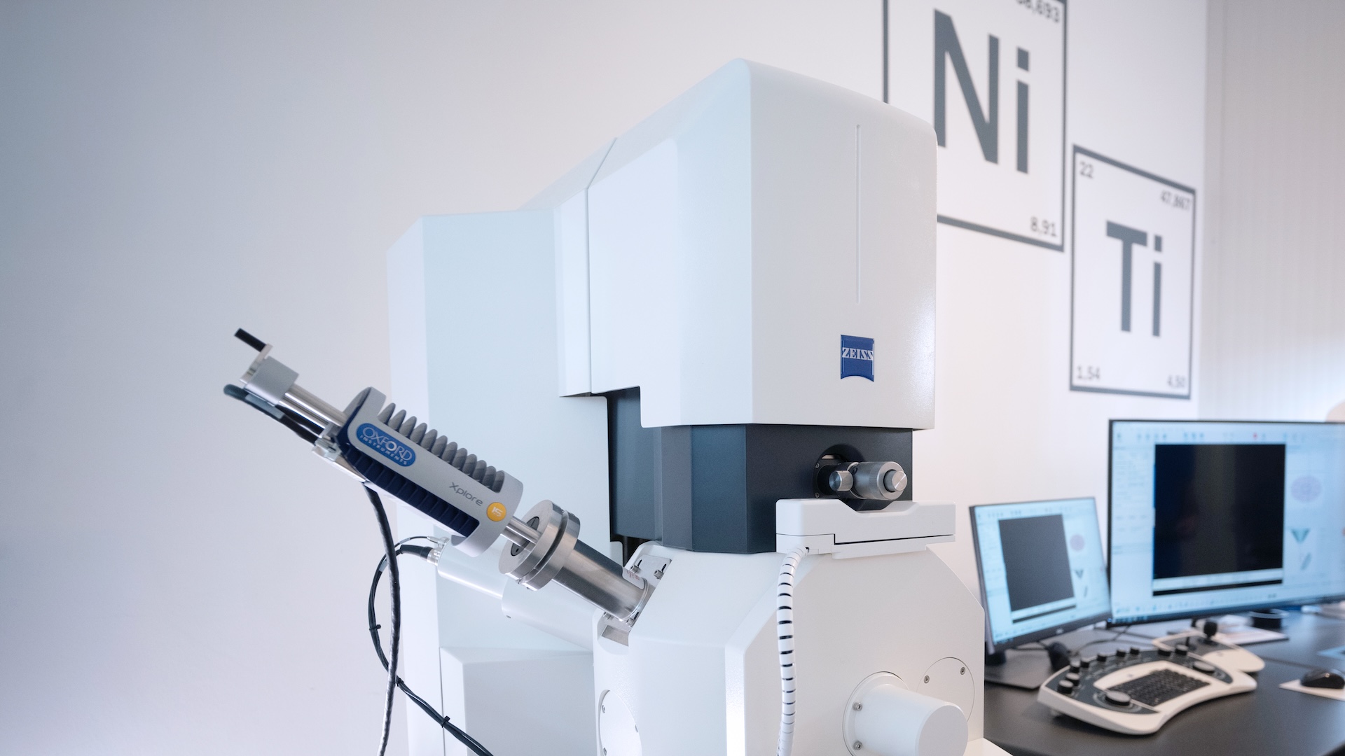 A ZEISS electron microscope setup in a laboratory environment with surrounding computer workstations and elemental wall charts.