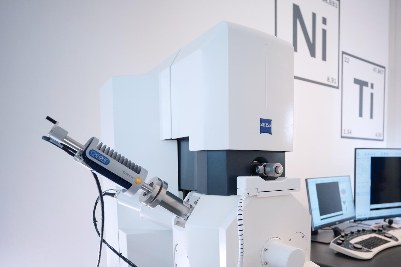 A ZEISS electron microscope setup in a laboratory environment with surrounding computer workstations and elemental wall charts.