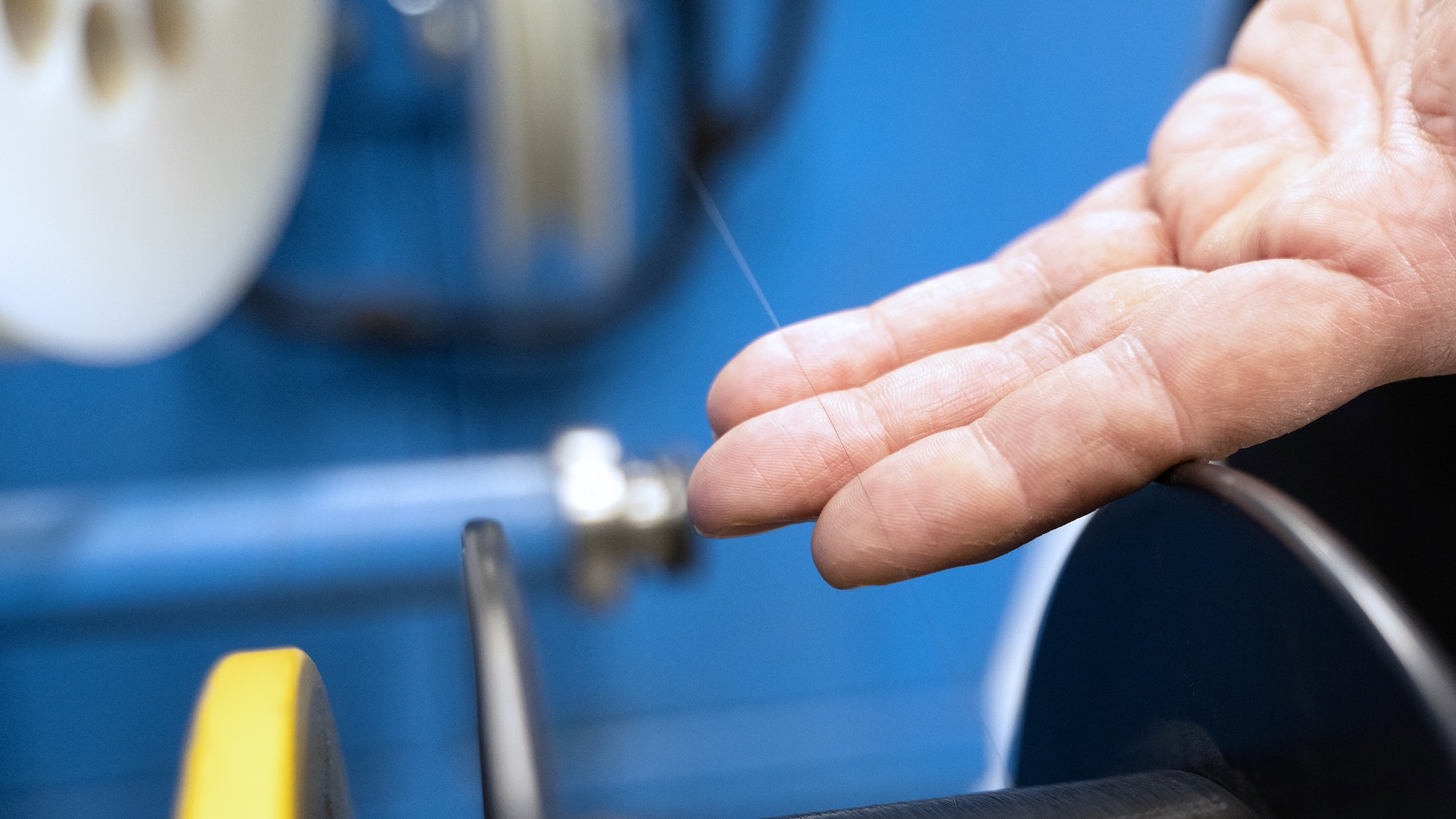 A close-up of a hand inspecting a fine optical fiber strand being processed on a precision machine in a ZEISS production environment.