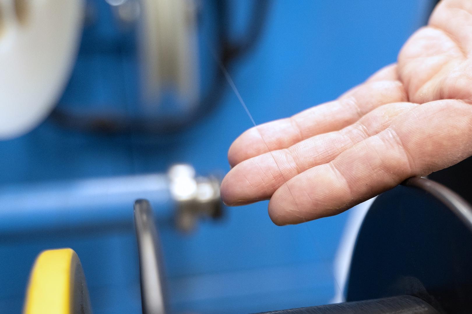 A close-up of a hand inspecting a fine optical fiber strand being processed on a precision machine in a ZEISS production environment.
