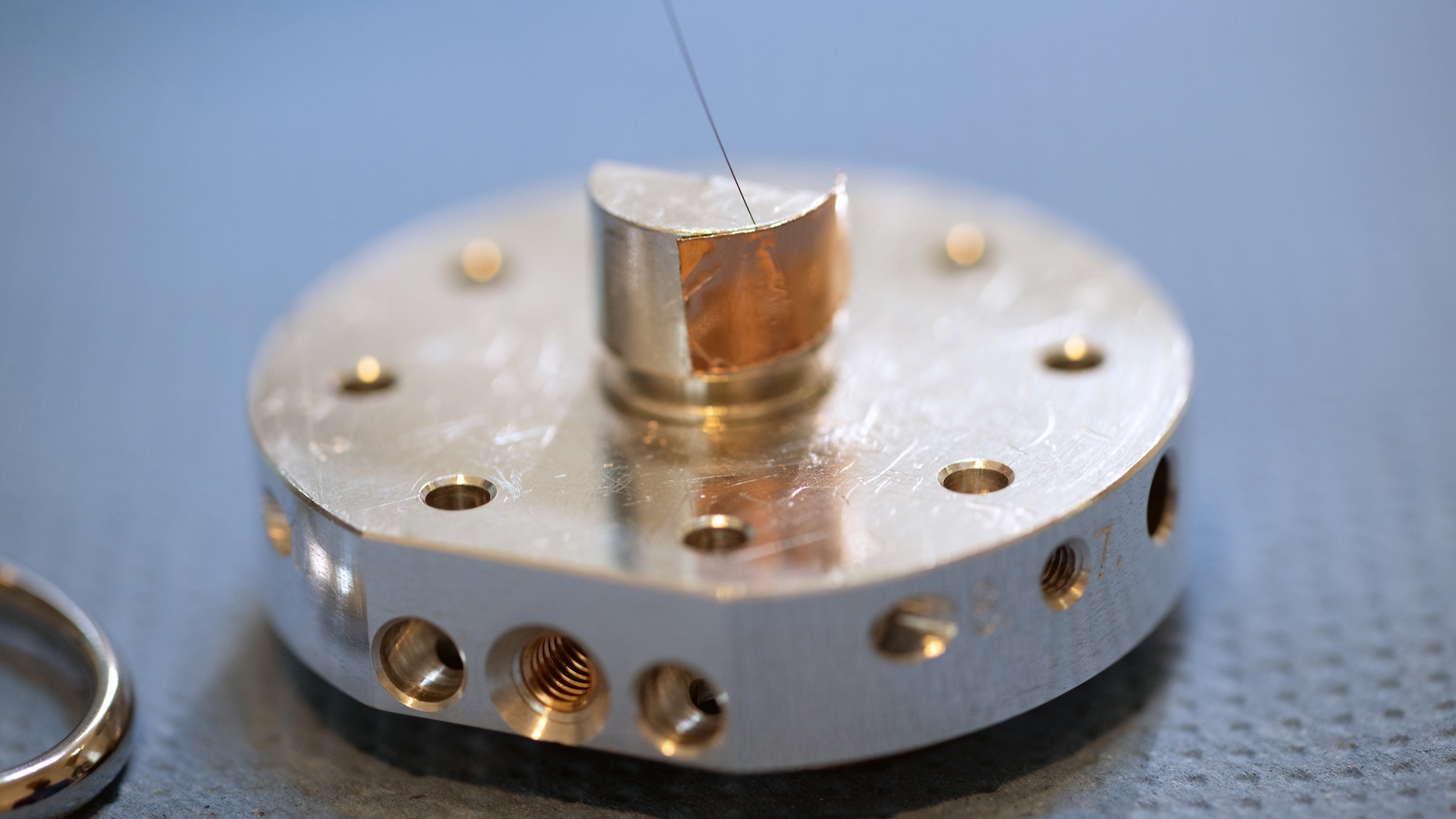 Close-up of a precision-engineered metallic component with multiple drilled holes and a thin wire, displayed on a textured surface, for ZEISS.