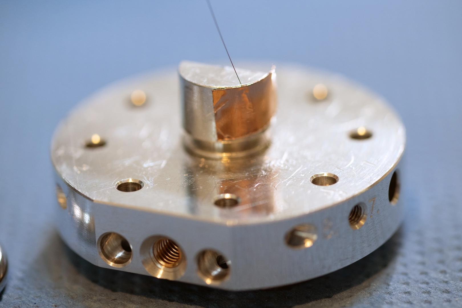 Close-up of a precision-engineered metallic component with multiple drilled holes and a thin wire, displayed on a textured surface, for ZEISS.