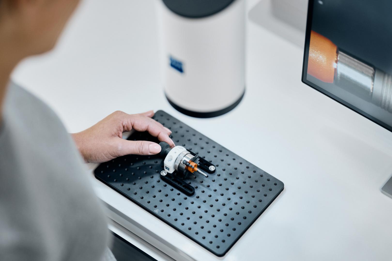 A person operates a ZEISS Smartzoom 100 to inspect a small mechanical component on a perforated work surface next to a monitor displaying analysis data.