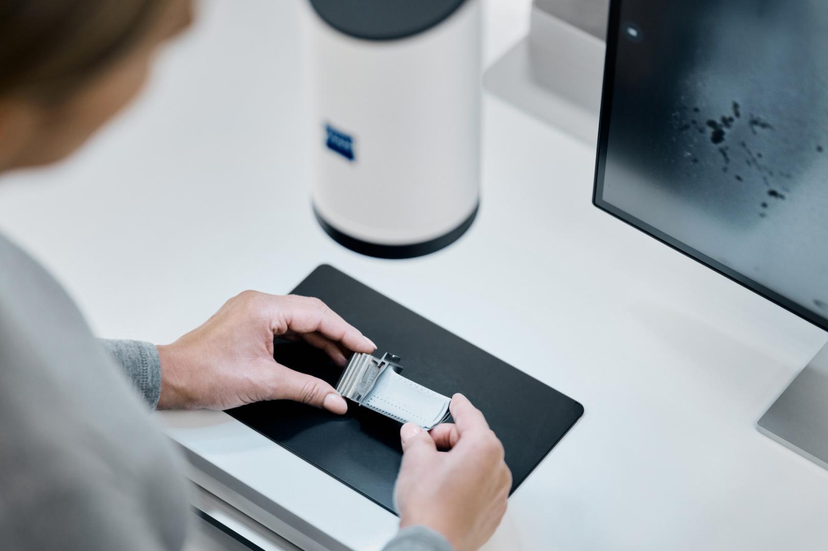 A person handling a microscope slide next to a monitor and ZEISS laboratory equipment on a clean workspace.