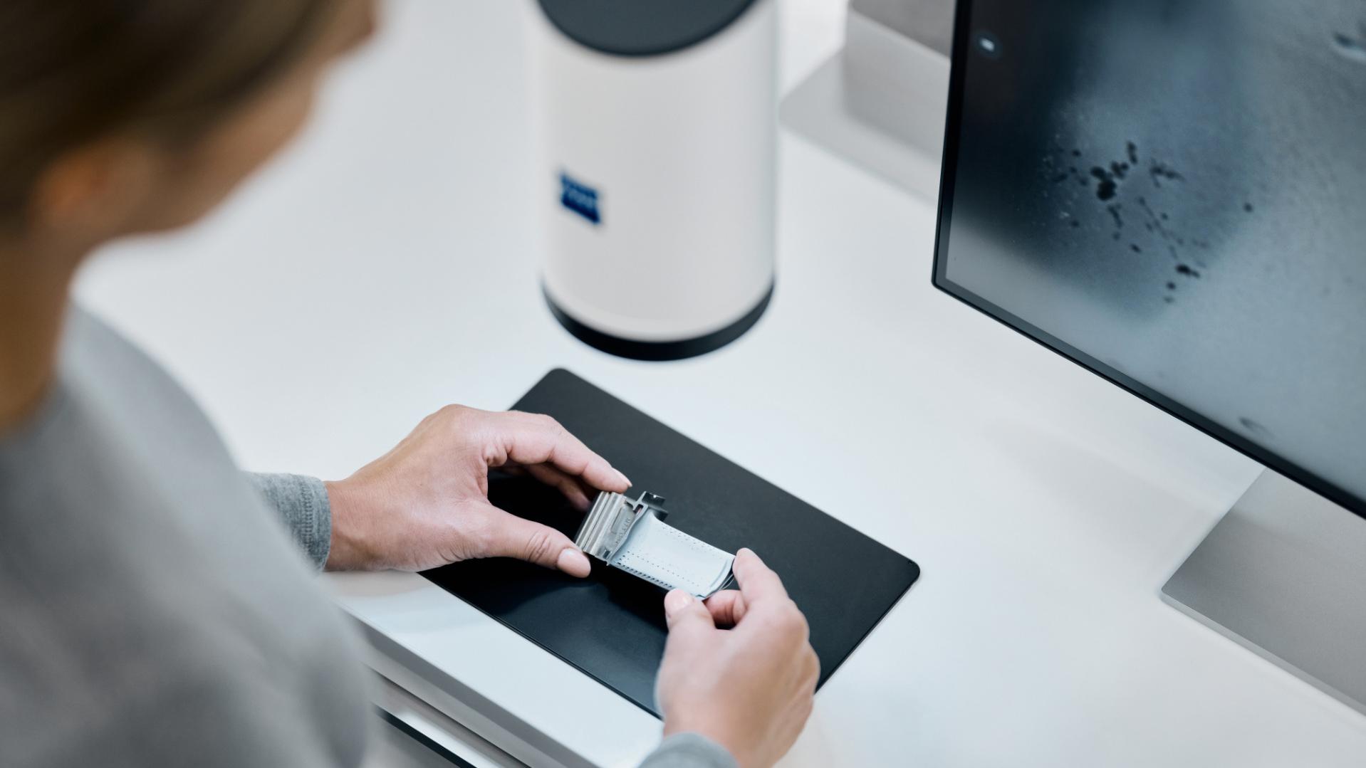 A person handling a microscope slide next to a monitor and ZEISS Smartzoom 100 on a clean workspace.