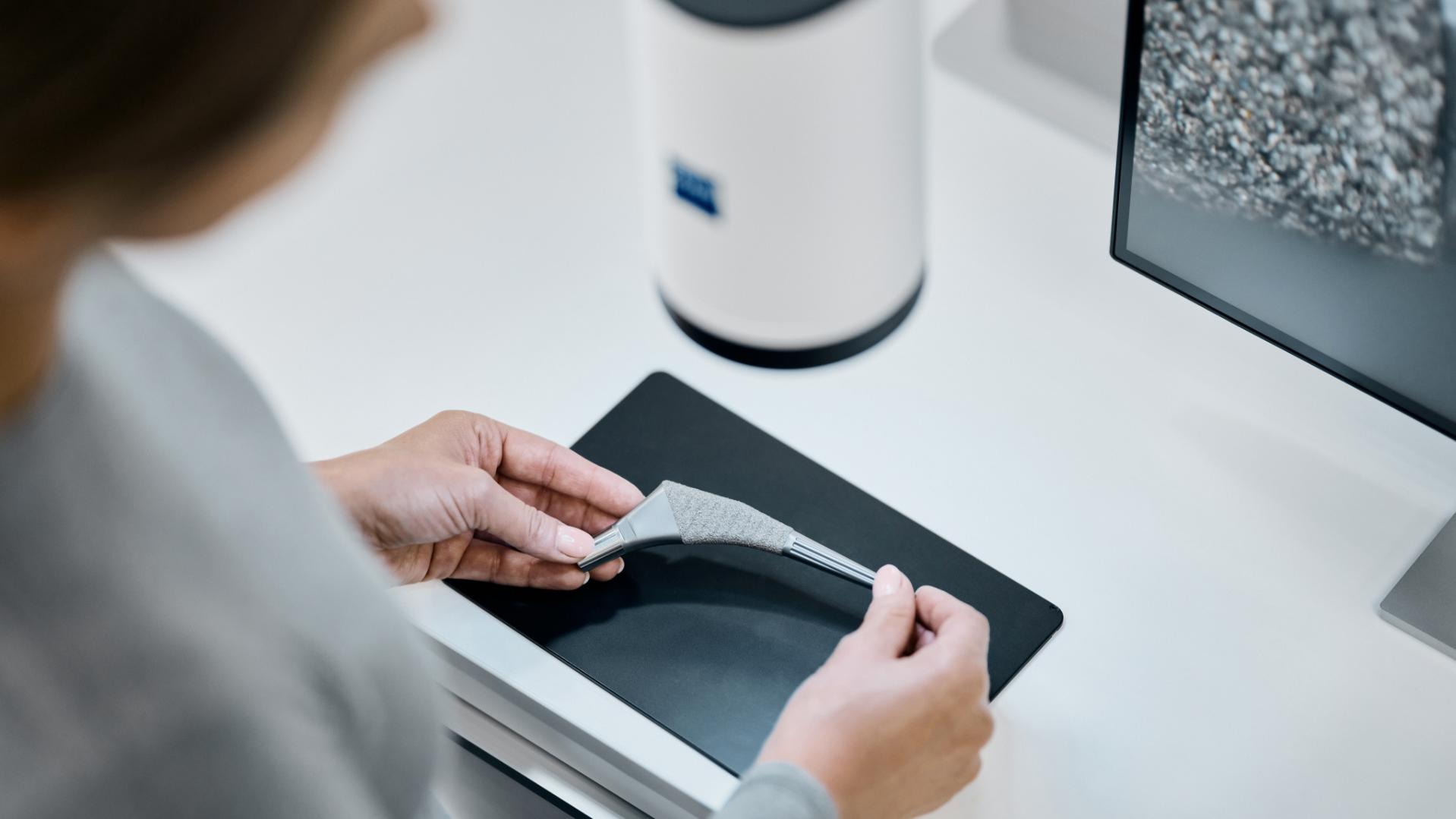 A person holding a 3D-printed part on a black surface next to a ZEISS device and a monitor displaying a magnified material structure.