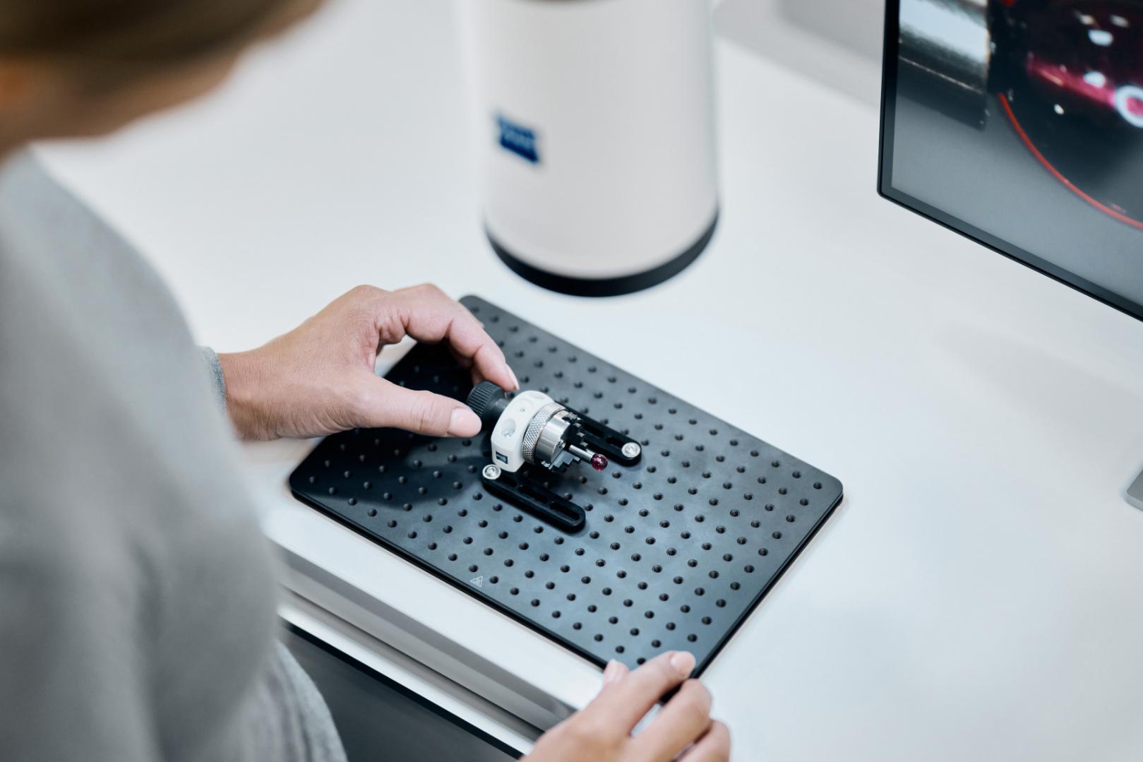 A person is positioning a mechanical component on a perforated base plate next to a ZEISS device and a computer monitor displaying a measurement interface.