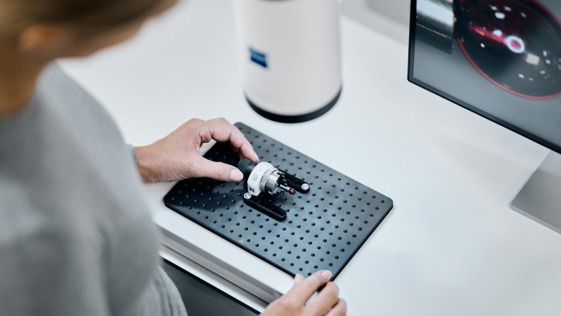 A person is positioning a mechanical component on a perforated base plate next to a ZEISS device and a computer monitor displaying a measurement interface.