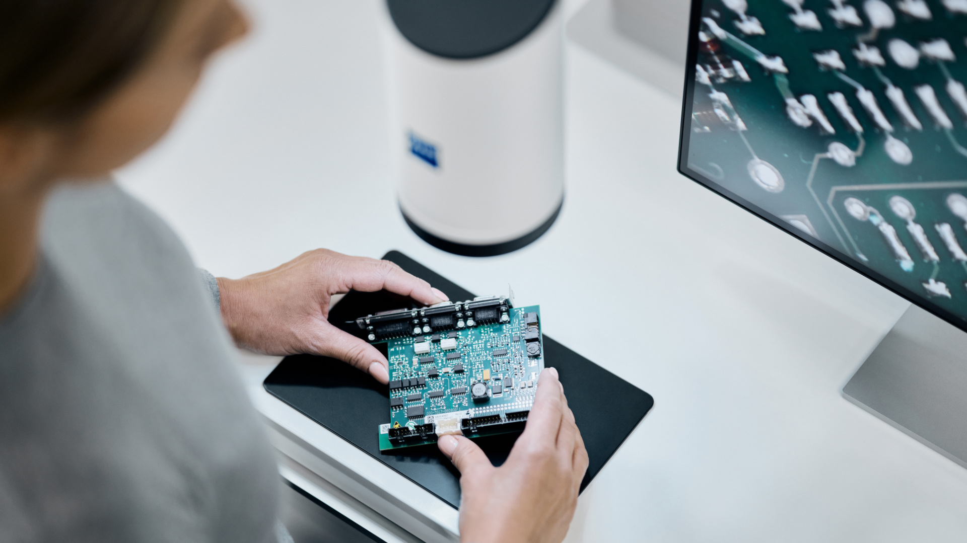 A person examines a circuit board in front of ZEISS Smartzoom 100 and a monitor displaying a magnified image of electronic components.