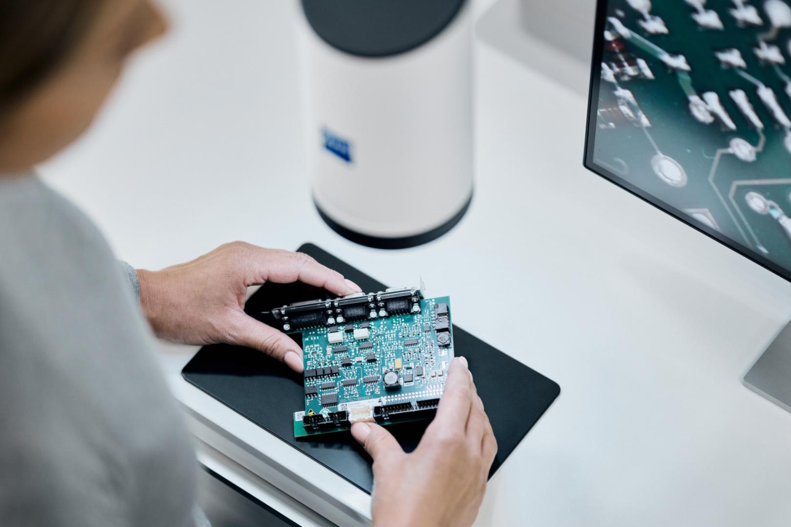 A person examines a circuit board in front of a ZEISS inspection device and a monitor displaying a magnified image of electronic components.