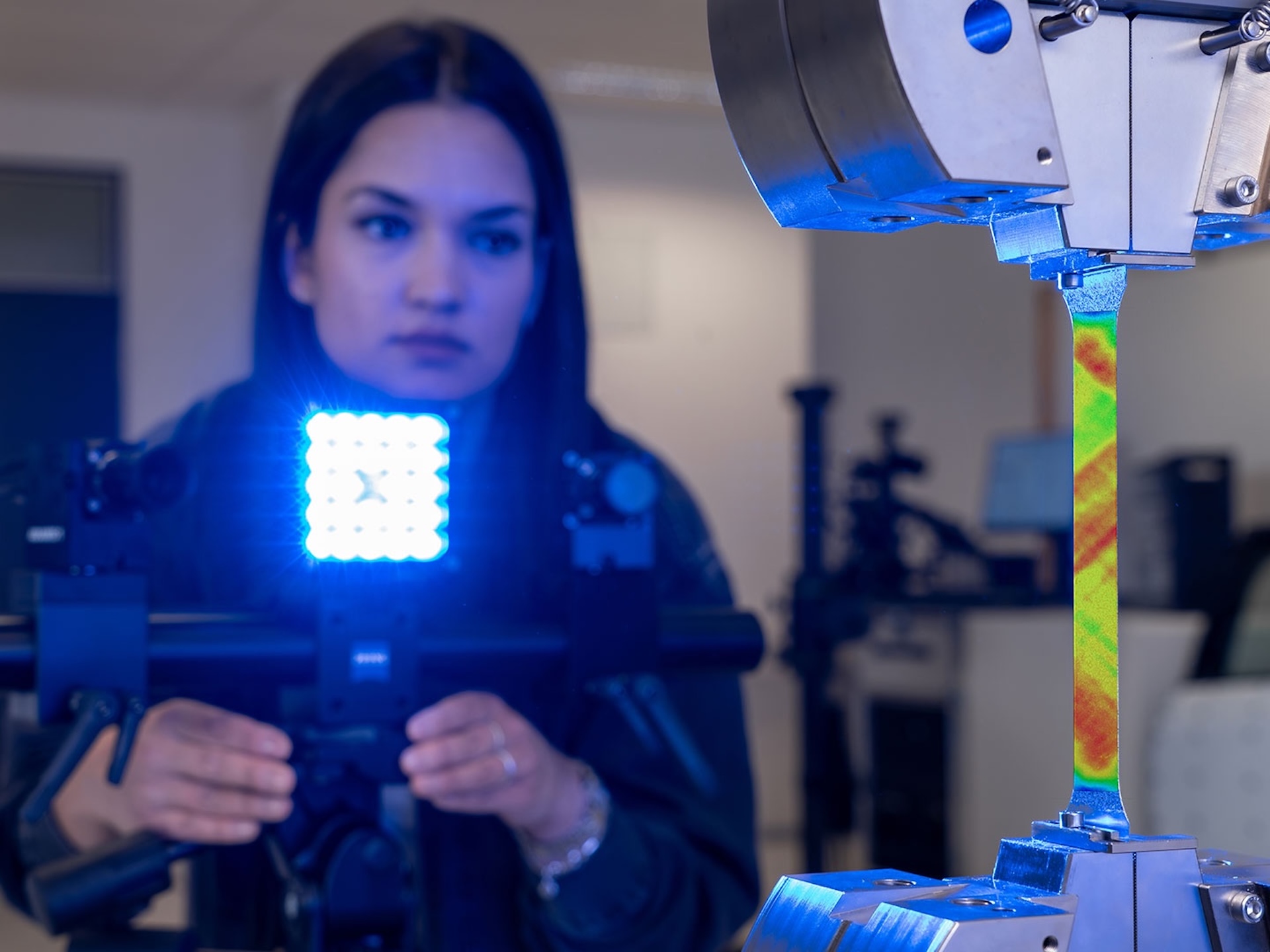 A woman operates a ZEISS optical measurement system analyzing material deformation under blue LED illumination in a laboratory setting.