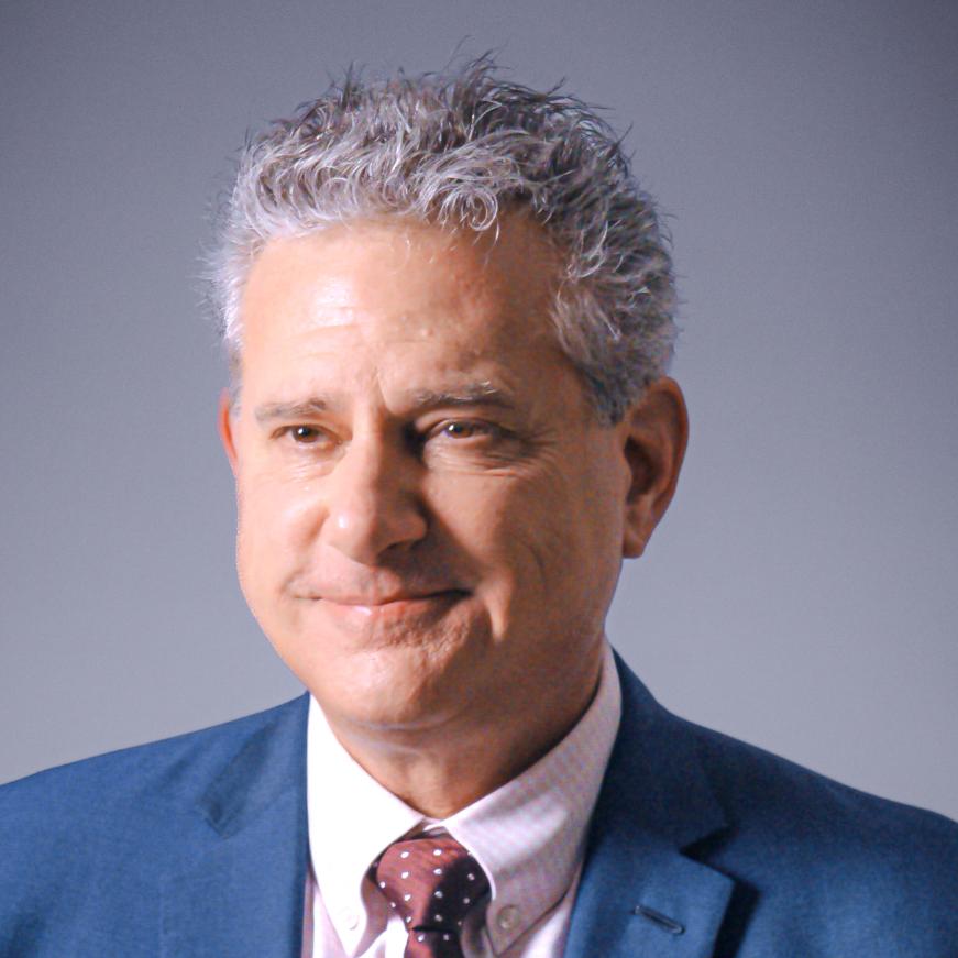 A middle-aged man with gray curly hair wearing a blue suit, white shirt, and red polka-dot tie, smiling slightly against a plain gray background.