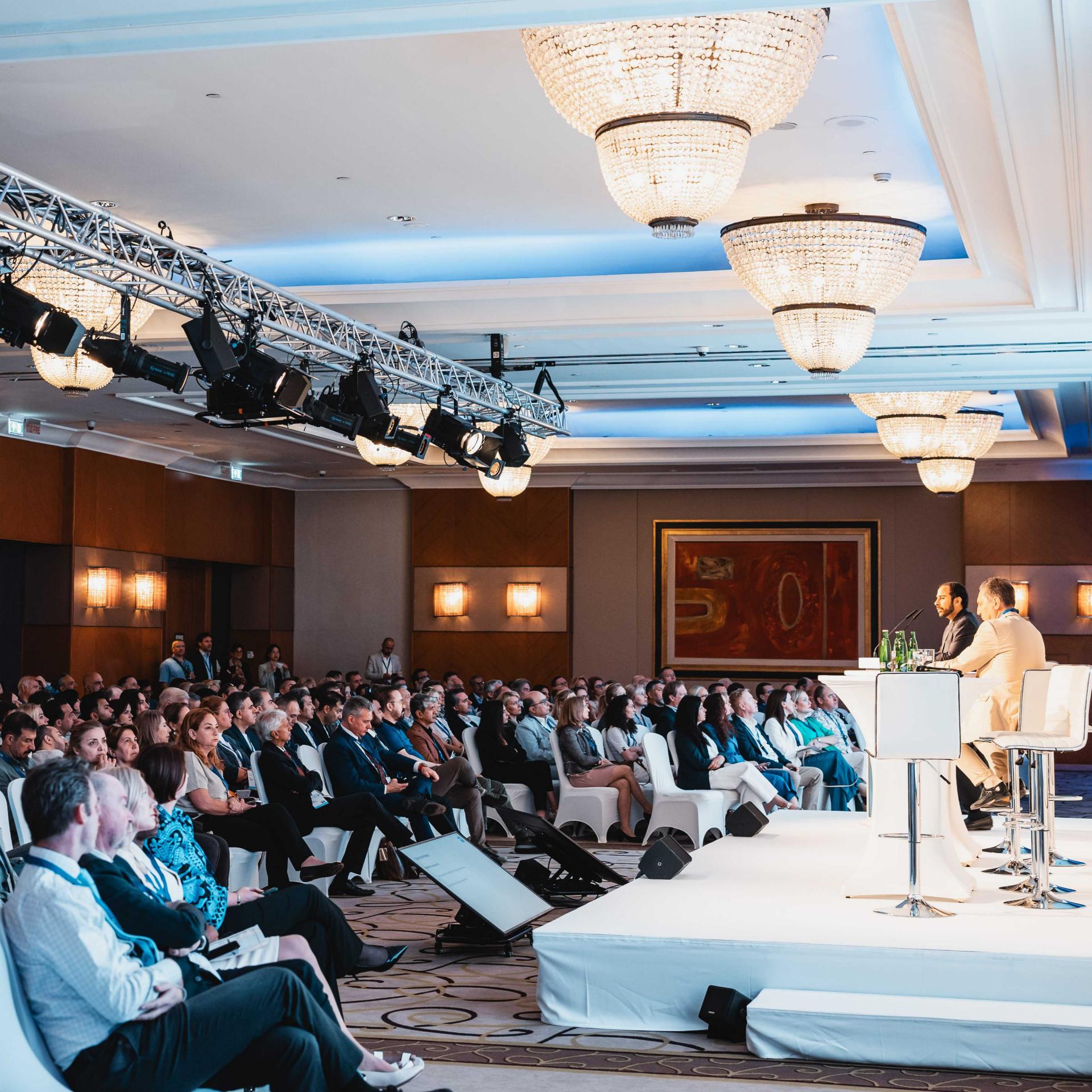 A large audience attentively listens to a panel discussion on a brightly lit stage at a professional ZEISS conference event.