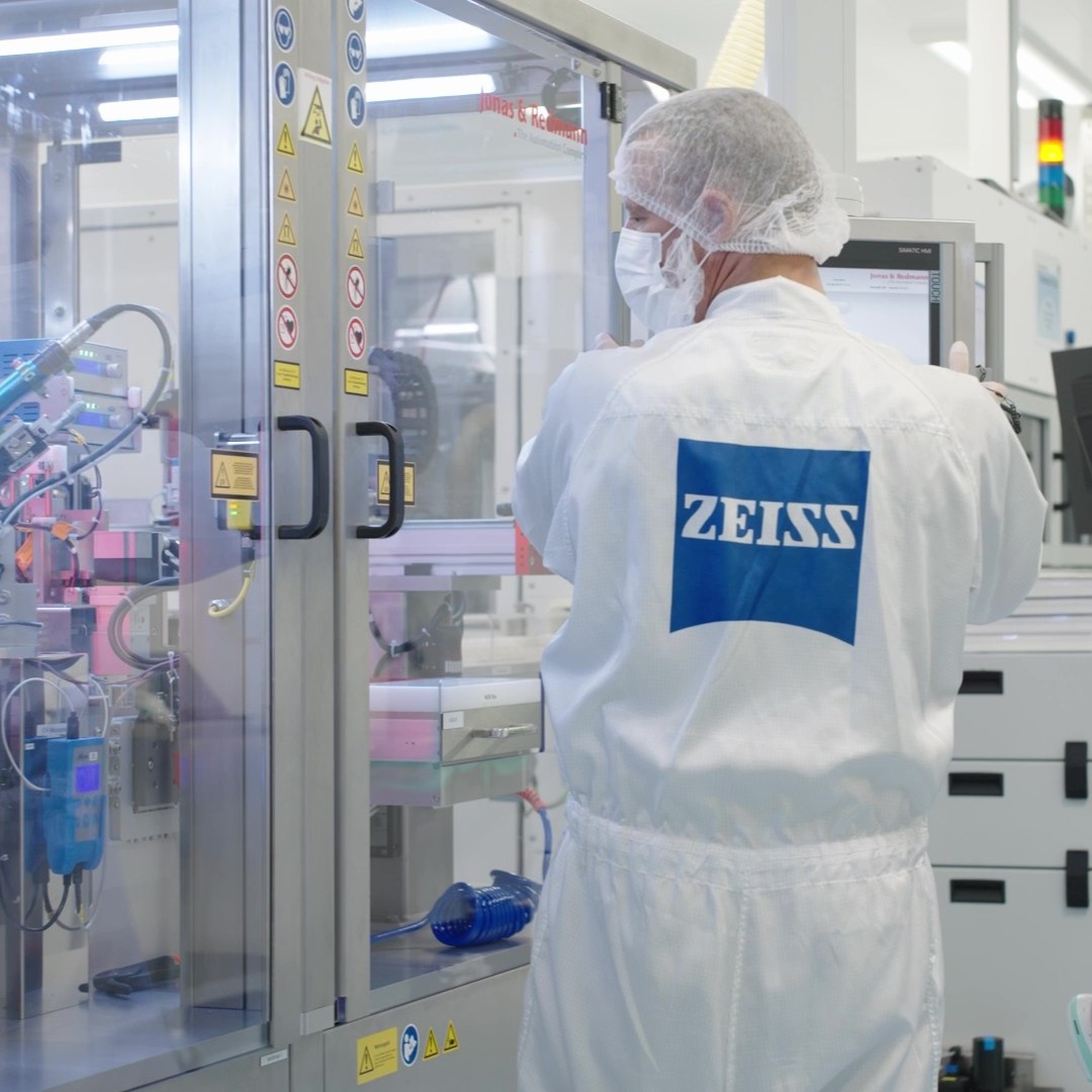 A ZEISS employee in cleanroom attire operates high-precision equipment inside a modern manufacturing facility.
