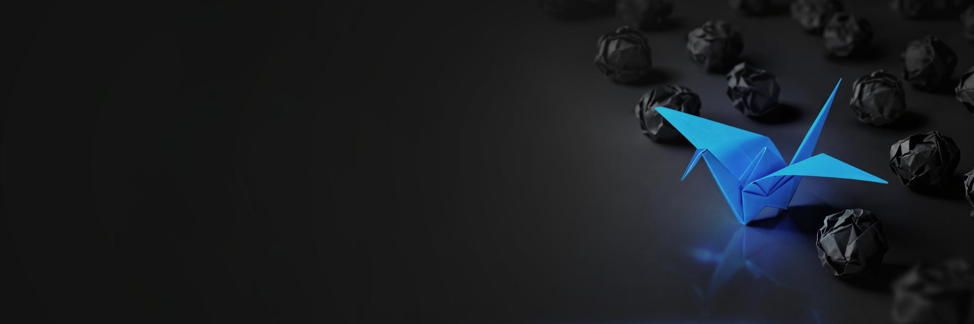 A single bright blue origami crane stands out among multiple black crumpled paper balls on a dark surface.