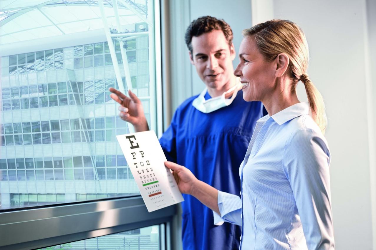 A woman holds an eye chart while standing next to a man in medical clothing near a large window, with a modern building visible outside.