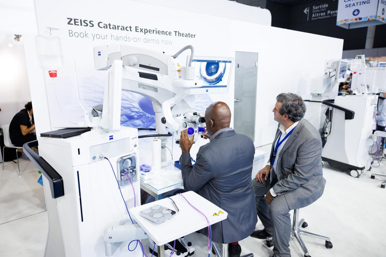 Two men in suits interact with advanced ophthalmic equipment at the ZEISS Cataract Experience Theater demonstration area.