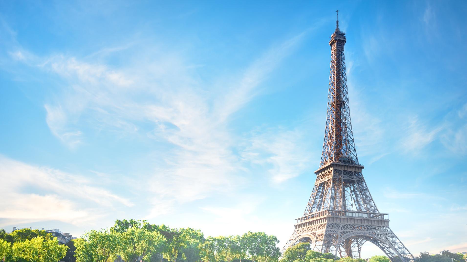 The image shows the Eiffel Tower rising above green trees under a bright blue sky with wispy clouds.