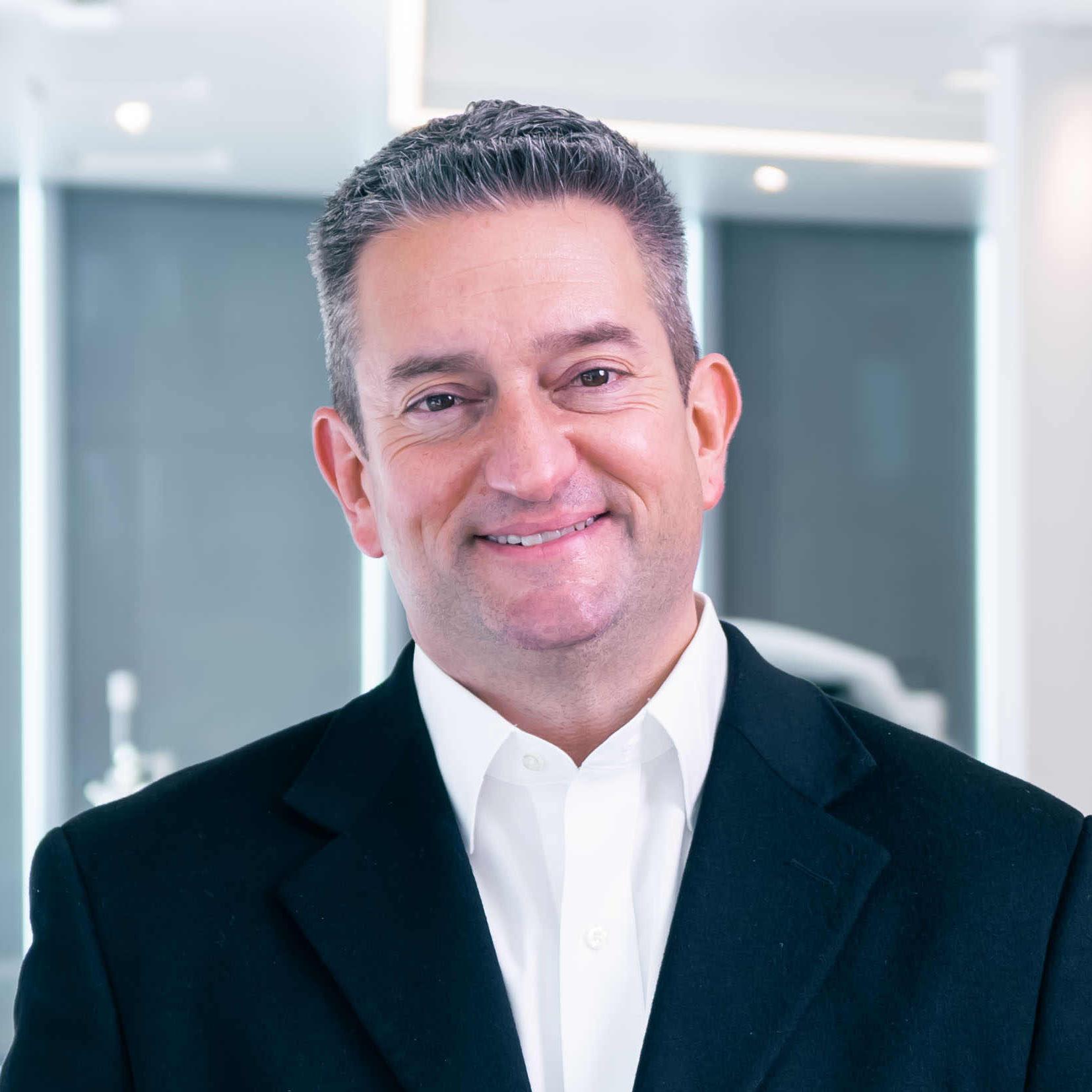 A professionally dressed man stands smiling with open hands in a modern, high-tech ZEISS medical or optical equipment showroom.