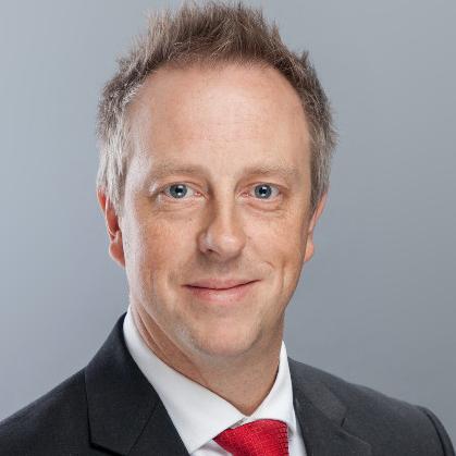 Professionelles Porträt eines lächelnden Manns mit schwarzem Anzug, weißem Hemd und roter Krawatte vor einem schlichten grauen Hintergrund, der ZEISS repräsentiert.