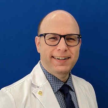 A smiling male professional wearing glasses, a white lab coat, and a dress shirt with tie stands in front of a solid blue background.