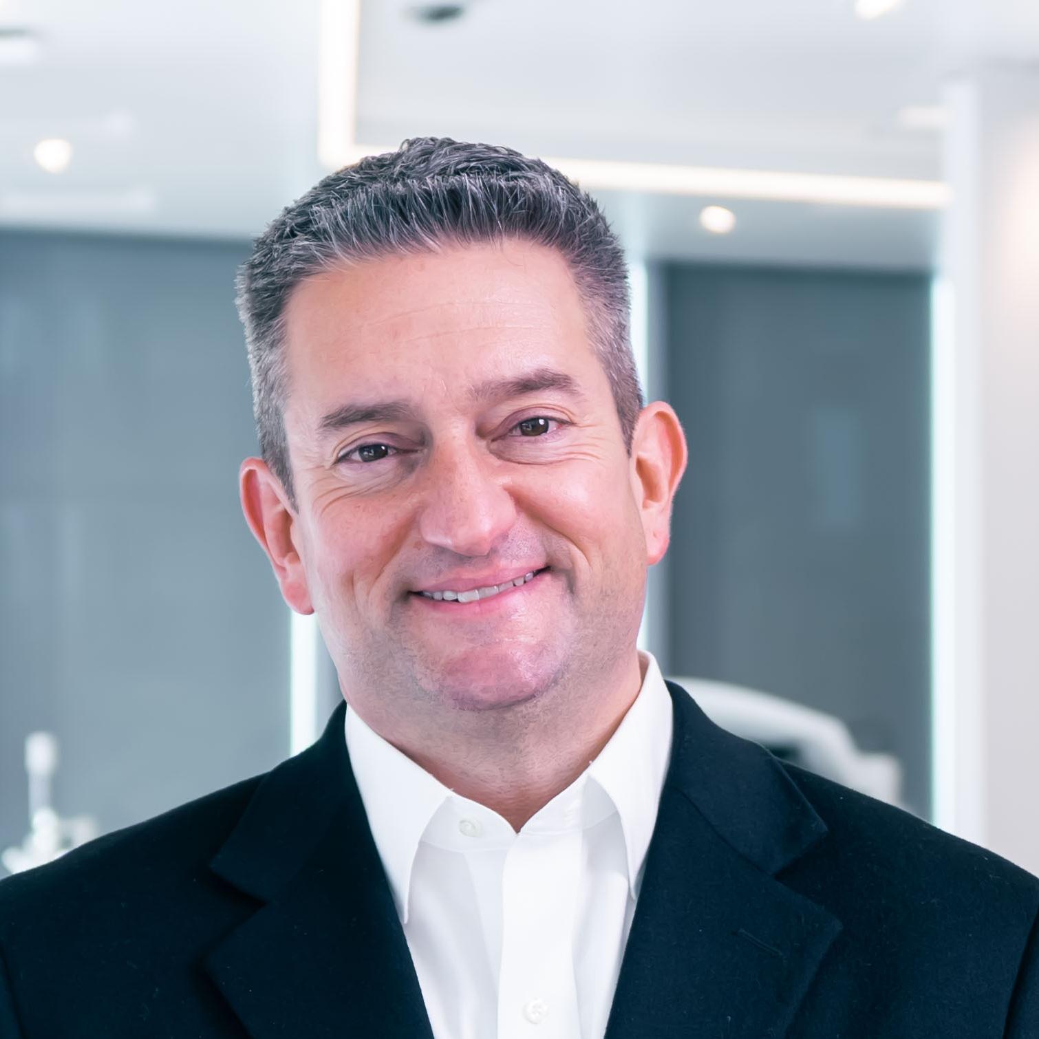 A professionally dressed man stands smiling with open hands in a modern, high-tech ZEISS medical or optical equipment showroom.
