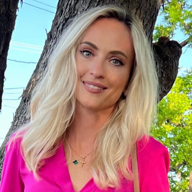 A woman with long blonde hair wearing a bright pink blouse stands in front of a tree with green foliage in the background.