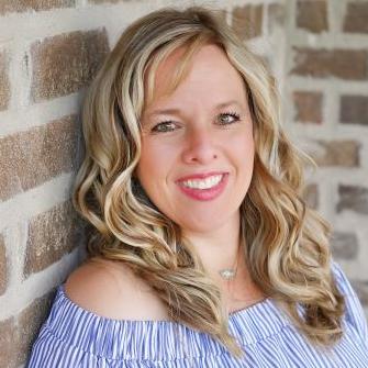 A woman with long blonde curly hair wearing a blue and white striped off-the-shoulder top smiles while standing in front of a brick wall.