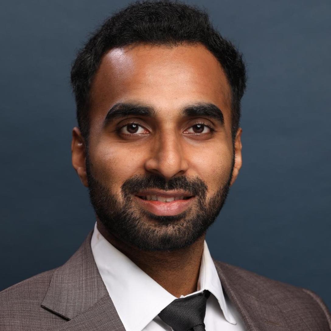 Dr. med. Aadithreya Varman dressed in a sharp brown suit, white shirt, and black tie against a dark blue background.