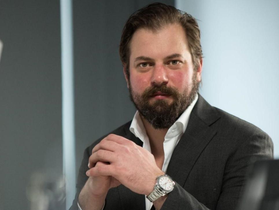 Dr. Florian Kretz in a dark suit sits at a desk in a modern office, hands clasped and wearing a silver wristwatch, with computer equipment in the foreground.