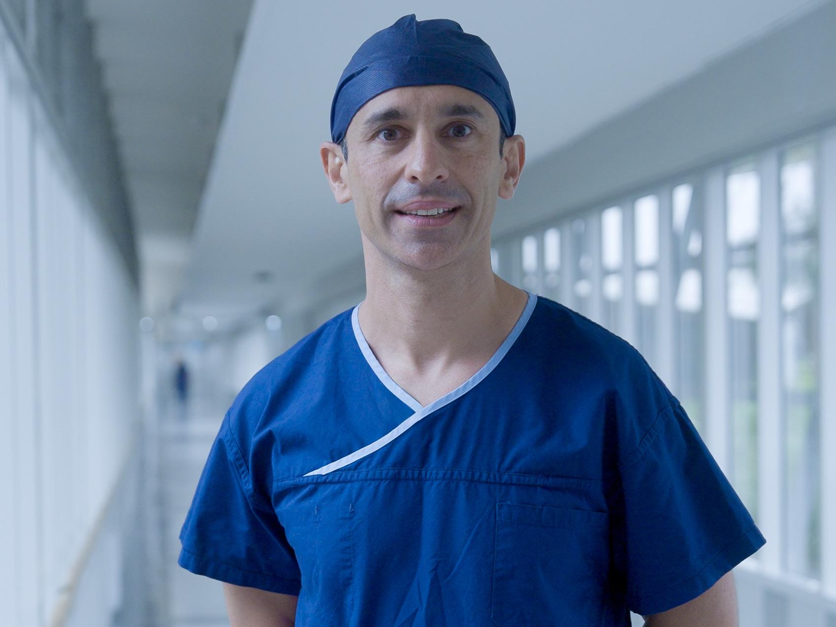 Prof. Kris Rallah-Baker wearing blue scrubs and a surgical cap stands in a bright hospital corridor.