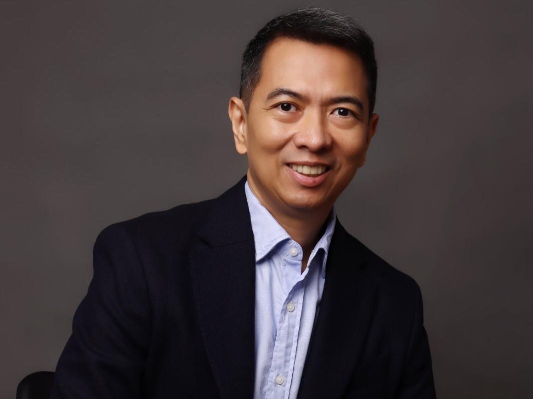 Dr. Manolette Roque is wearing a navy blazer, light blue button-down shirt in front of the gray studio background.