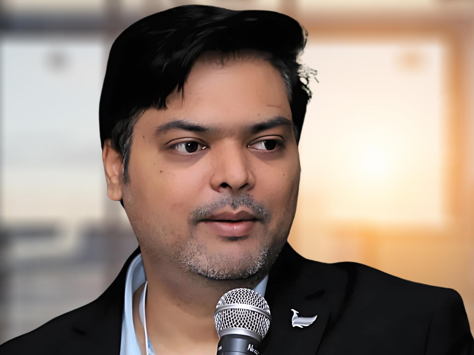 Dr. Nilesh Kumar is in a black blazer holding a microphone in a bright, modern office setting.