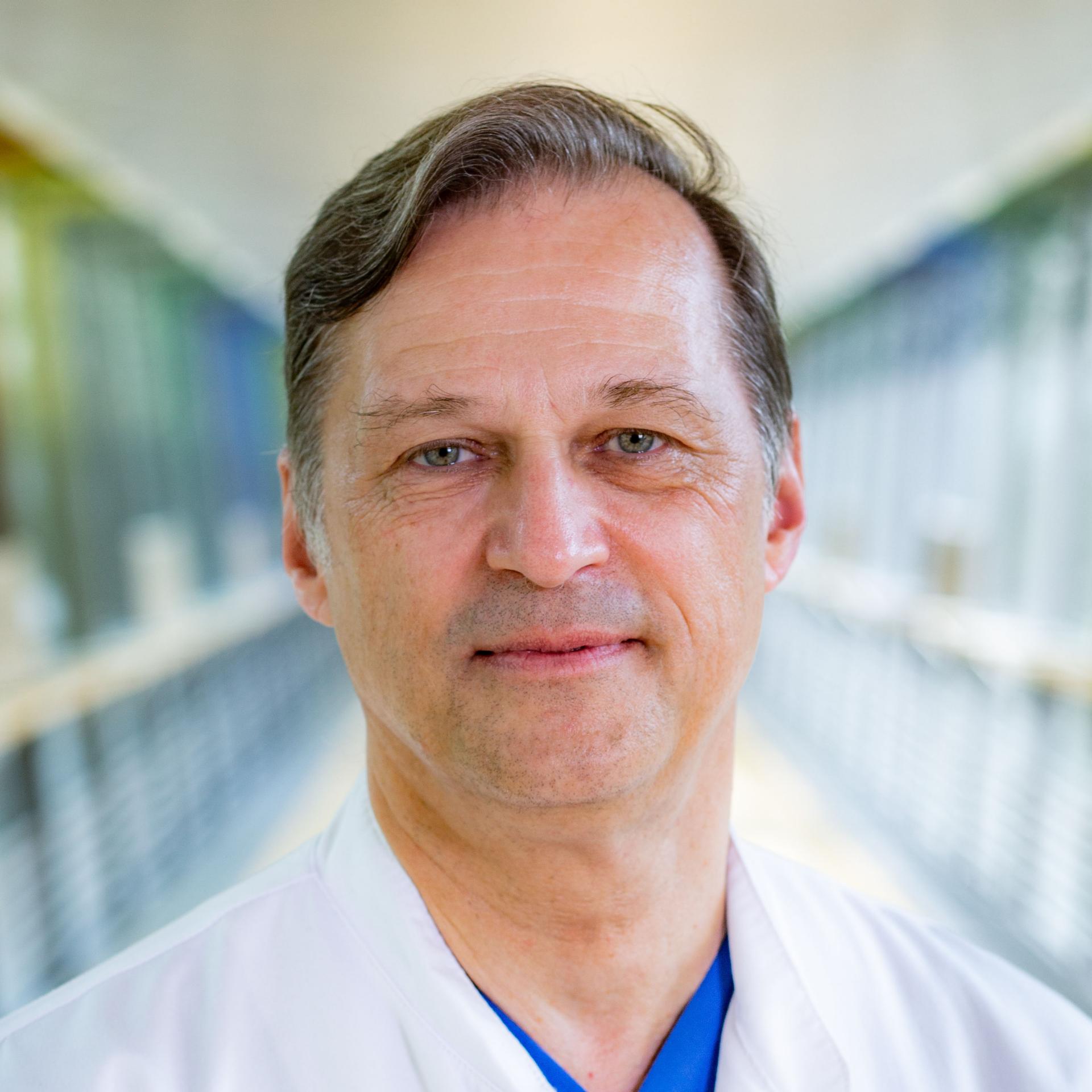 Un médecin d'âge moyen, vêtu d'une blouse blanche et d'une tenue chirurgicale bleue, se tient dans un couloir bien éclairé.