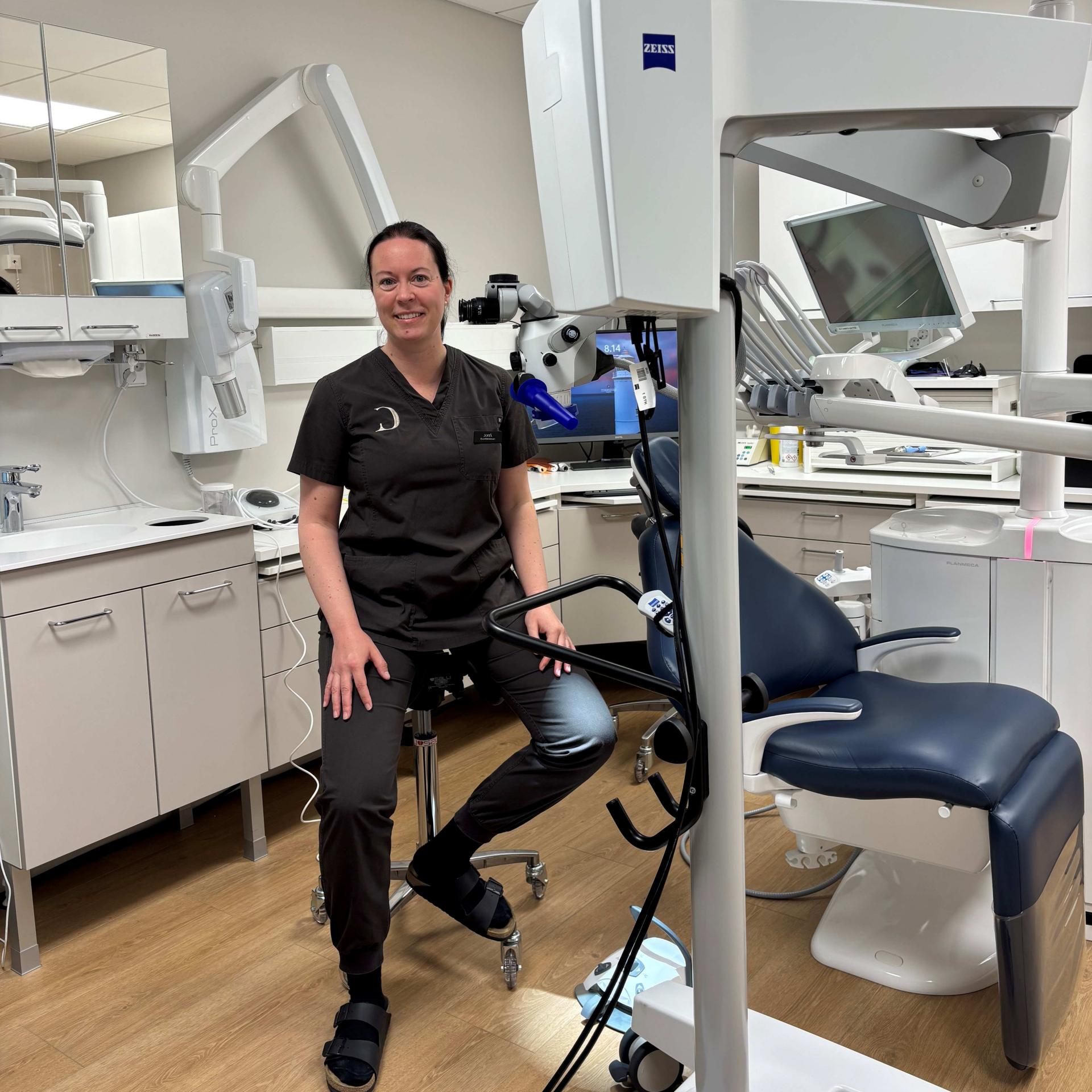 A dental professional sits in a modern clinic beside a ZEISS medical microscope and examination chair, surrounded by advanced diagnostic equipment.