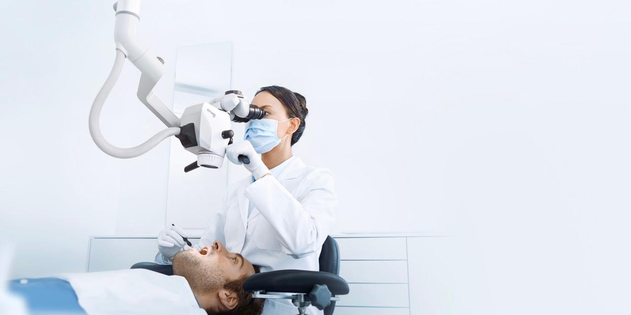 A dentist examines a patient using a ZEISS dental microscope in a bright, modern clinic.