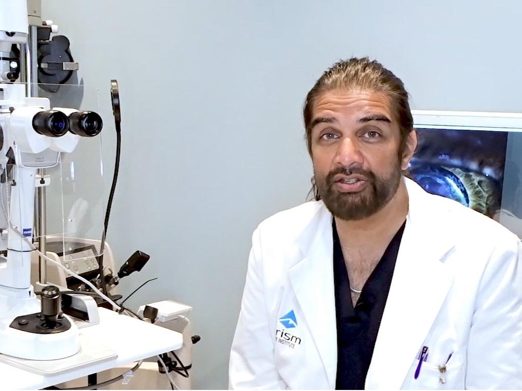 Ike Ahmed, MD in a white lab coat sits beside an ophthalmic examination device in a clinical setting, with an eye image displayed on a screen in the background.