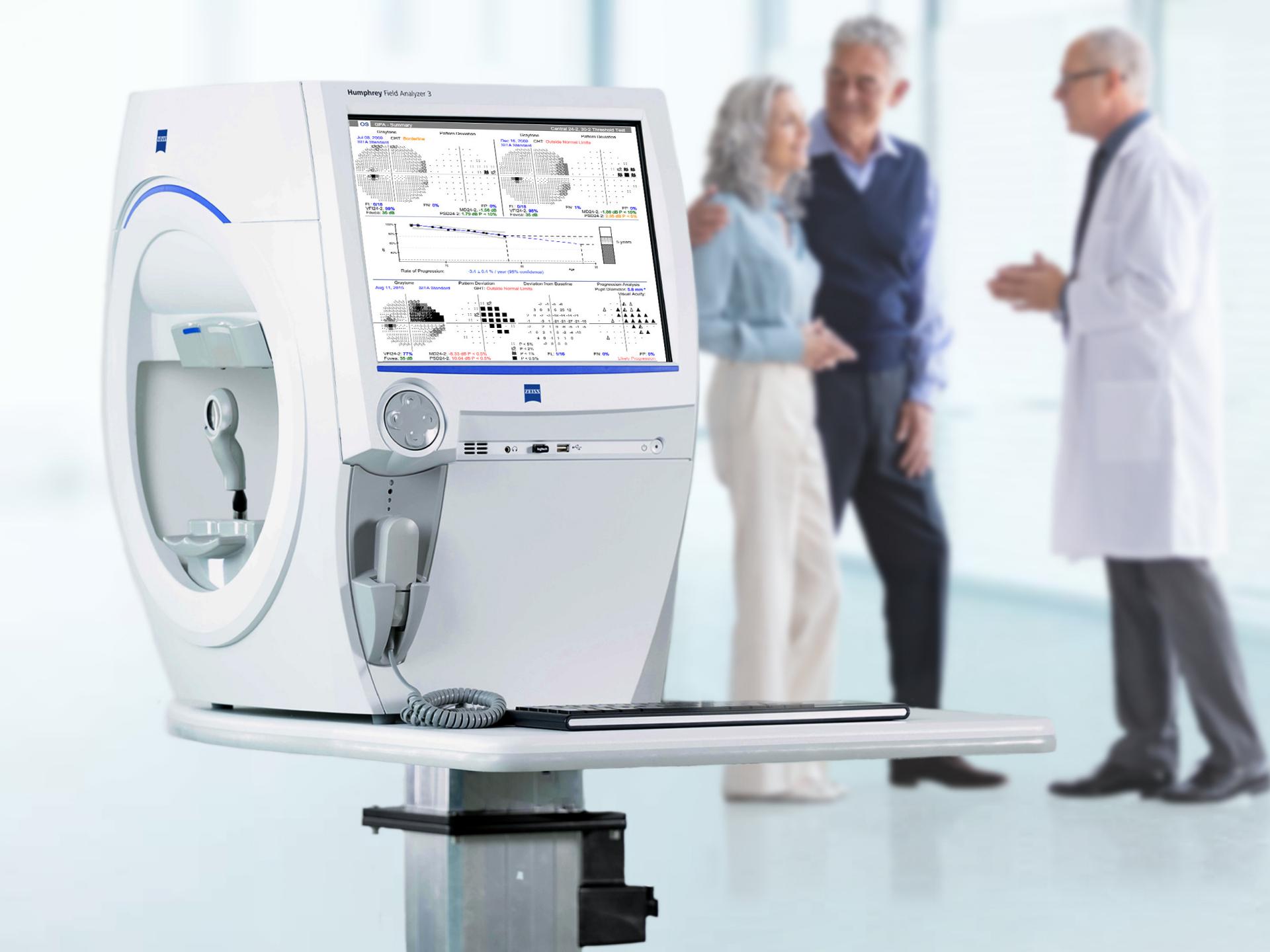ZEISS medical diagnostic device with a display screen showing test results, positioned in the foreground of a clinical setting with three people conversing in the background.