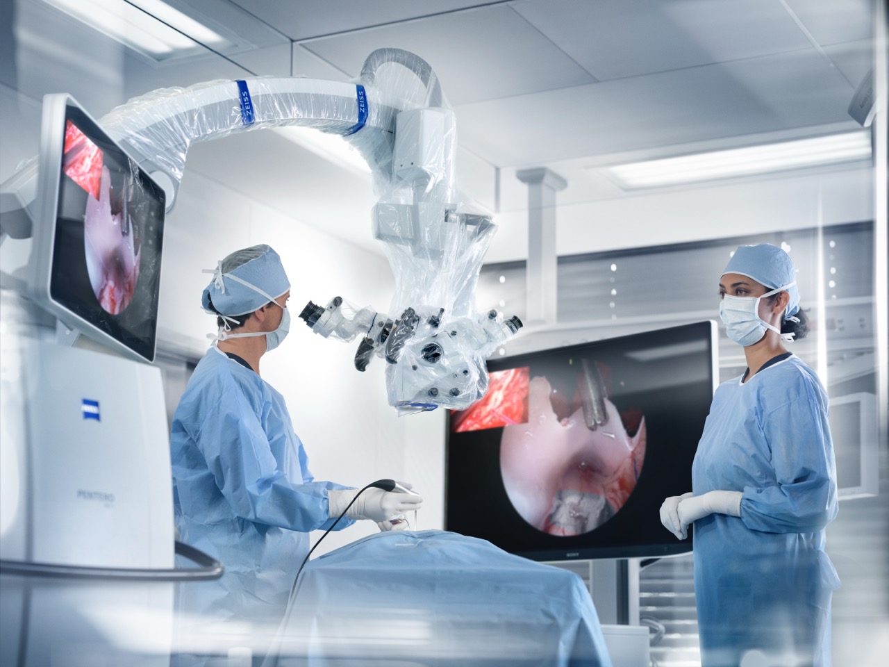 Two surgeons in an operating room use a ZEISS surgical microscope system while viewing high-resolution imagery on ZEISS monitors during a medical procedure.