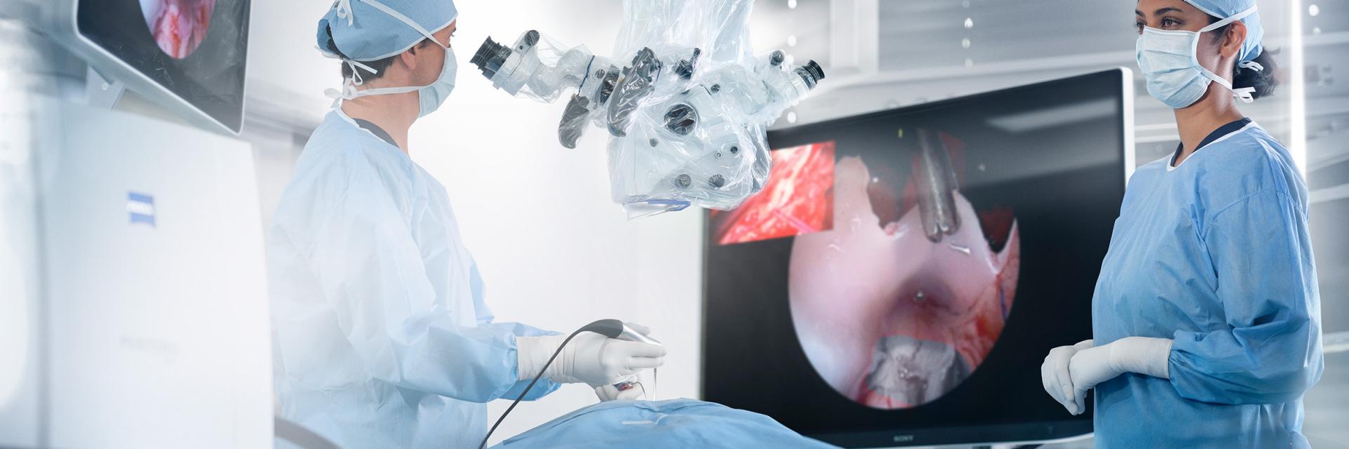 Two surgeons in an operating room use a ZEISS surgical microscope system while viewing high-resolution imagery on ZEISS monitors during a medical procedure.