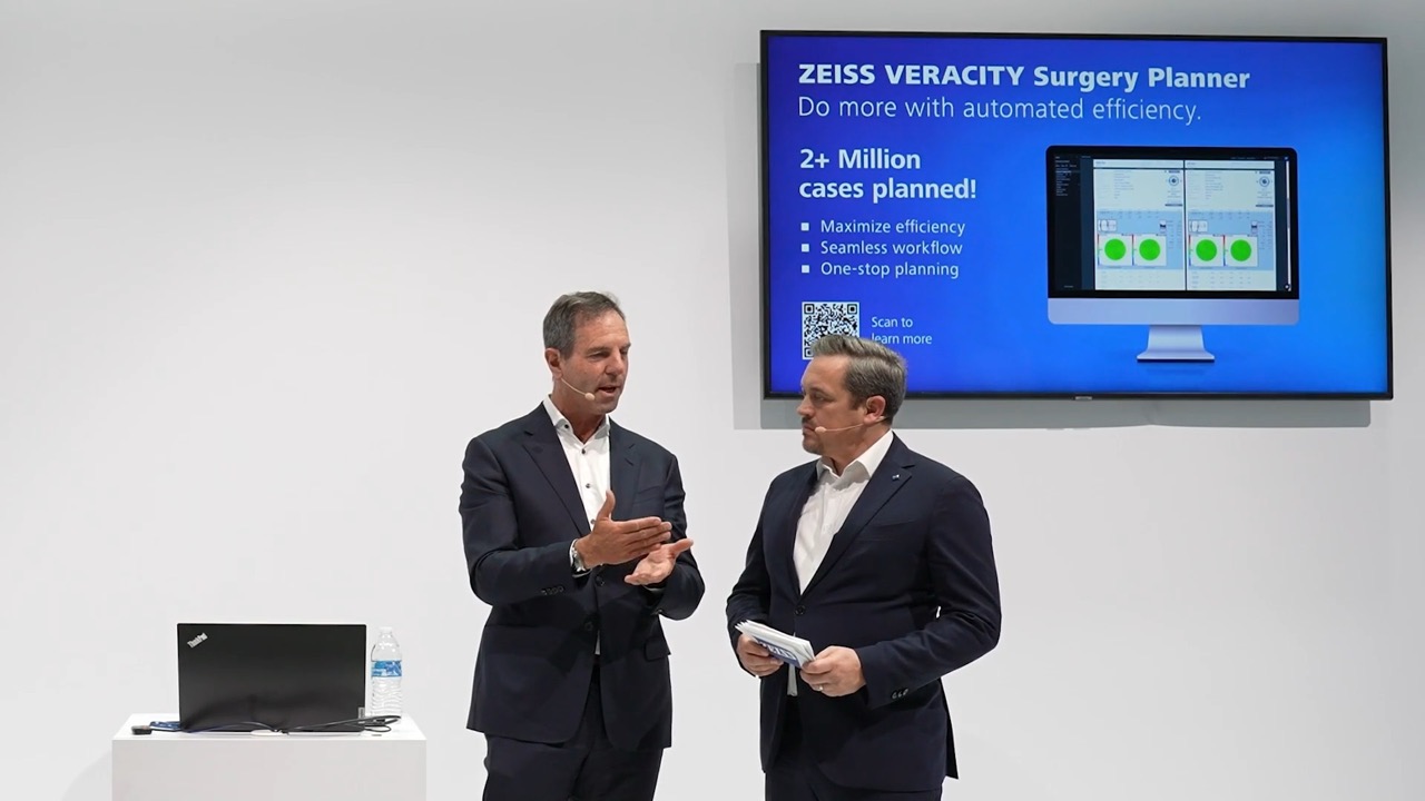Two men in business attire are speaking on stage beside a screen displaying information about the ZEISS VERACITY Surgery Planner and a laptop on a pedestal.
