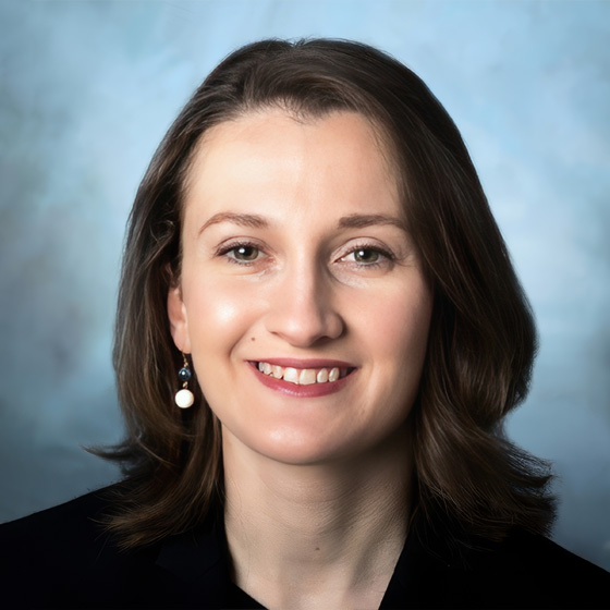 Portrait of a smiling woman with shoulder-length brown hair wearing a black top and pearl earrings, set against a soft blue background.